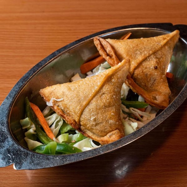 Two golden-brown fried samosas, triangular in shape, served in a metal dish with shredded vegetables.