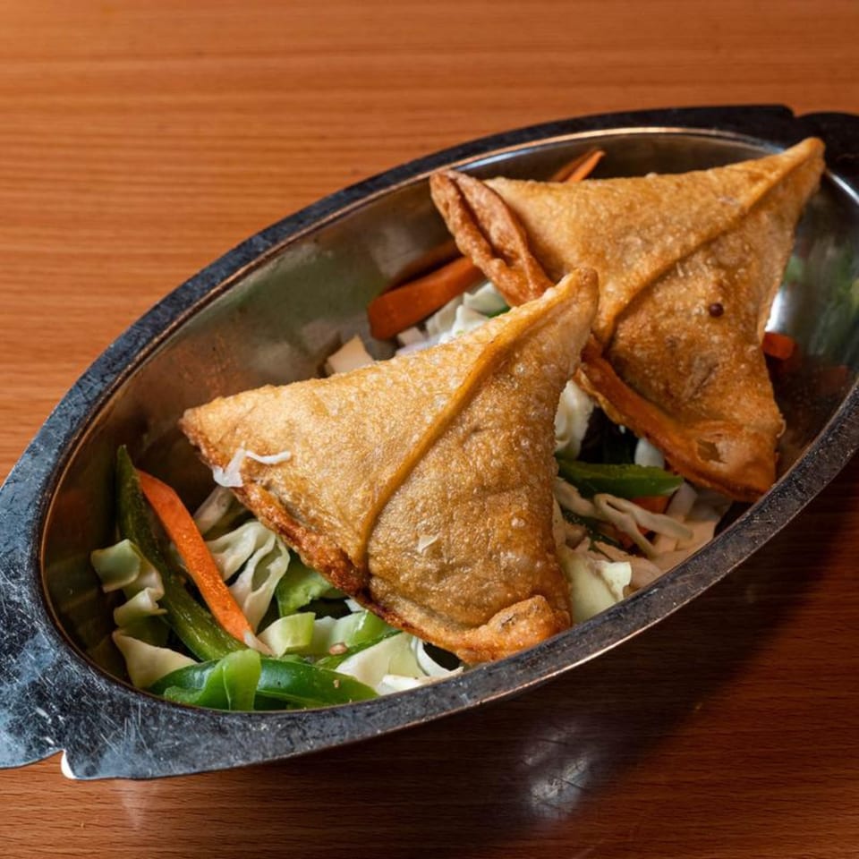 Two golden-brown fried samosas, triangular in shape, served in a metal dish with shredded vegetables.