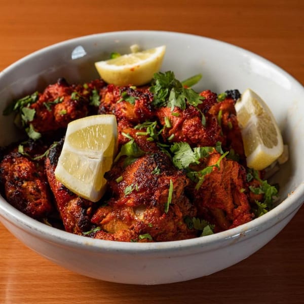 A white bowl filled with red tandoori chicken, garnished with fresh cilantro and lemon wedges.