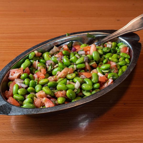 Edamame salad with diced tomatoes and red onions in a silver serving dish with a spoon.