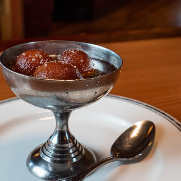 Three gulab jamun in a silver goblet with a spoon on a white plate, on a wooden table.