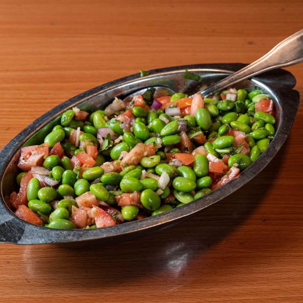 A metal dish filled with vibrant green edamame, diced tomatoes, and red onions, with a spoon.