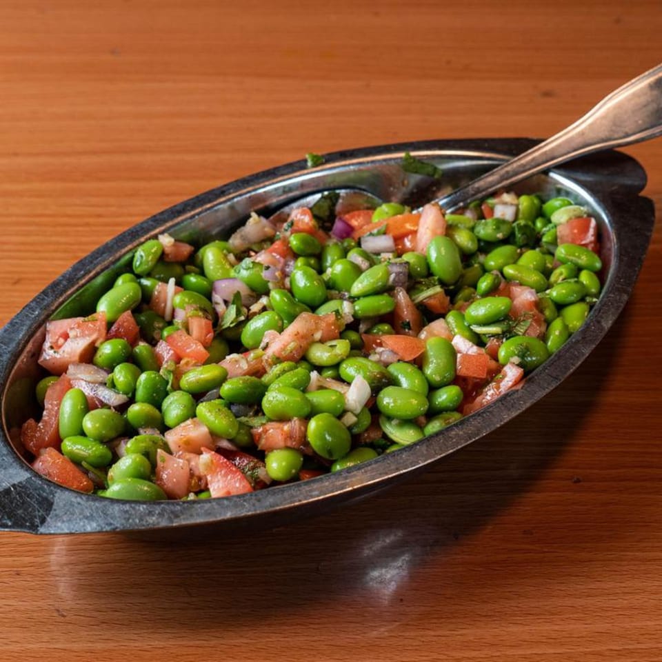 A metal dish filled with vibrant green edamame, diced tomatoes, and red onions, with a spoon.