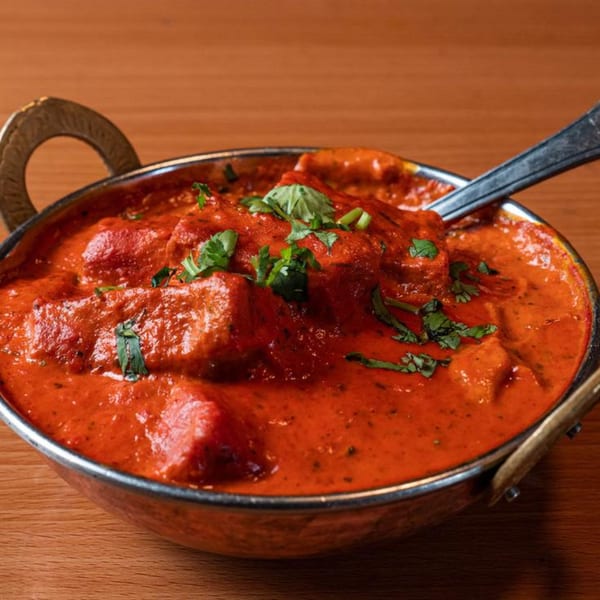 A close-up of a metal bowl filled with vibrant red curry, garnished with fresh green herbs.