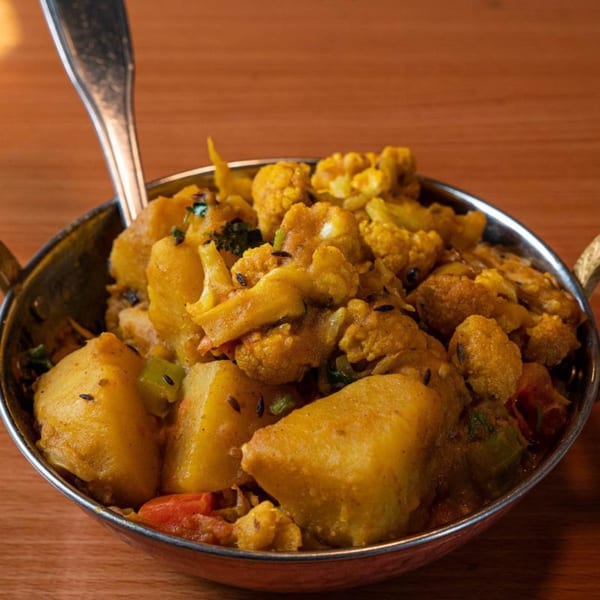 A metal bowl of Aloo Gobi, a Tibetan dish with potatoes and cauliflower, on a wooden table.